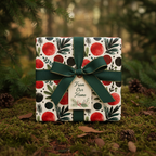Elegant Christmas gift wrap with red, green, and black botanical patterns, tied with a dark green ribbon and gold-edged bow, on a mossy forest floor.