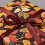 Close-up of autumn leaf floral wrapping paper on a square gift box with burgundy ribbon and bow.