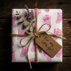 Square gift wrapped in baby girl wrapping paper with pink footprints, twine bow, lavender sprig, and tag.