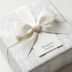Elegant baptism wrapping paper with gray angel and cross pattern, cream bow, and tag by Wrapped Studios.