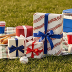 Baseball-themed birthday wrapping paper on gifts with red and blue ribbon, sitting on a picnic blanket with hotdogs.