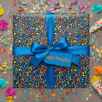Colorful celebration gift wrapped in confetti-patterned paper with a blue satin ribbon and "Happy Thoughts" tag.