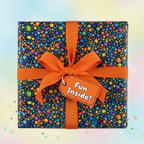 Square gift box wrapped in colorful confetti dot pattern wrapping paper with orange ribbon and "Fun Inside!" tag.