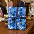 Blue graduation wrapping paper with caps and "Congrats!" text by Wrapped Studios, tied with a navy ribbon.