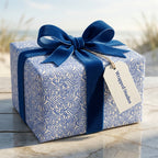 Elegant blue and white wavy abstract gift wrap with a thick velvet ribbon bow and Wrapped Studios tag on a beachside table.