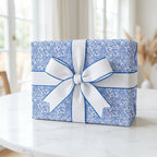 Modern blue and white abstract patterned gift wrap with a white grosgrain ribbon bow and blue trim, sitting on a marble table.