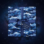 Square gift wrapped in navy star and cloud wrapping paper with a satin bow and tag on a space background.