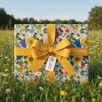 Square gift wrapped in floral paper with a gold ribbon, set in a sunny meadow.