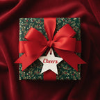 Festive Christmas gift wrapped in floral holiday wrapping paper with a red satin bow and "Cheers" tag, on red fabric.