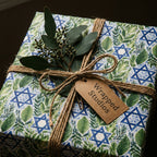Elegant Hanukkah gift wrap with blue Star of David and green foliage, tied with twine by Wrapped Studios.