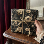 Gift wrapped in dark floral wrapping paper with gold ribbon, held in a hand with lace cuff.