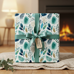 Luxury botanical wrapping paper with teal, blue, and gold leaf patterns, a green satin ribbon, and gift tag on a cozy blanket by a fireplace.