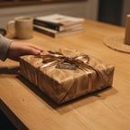 Hand reaches for a modern gift wrapped in tan paper with a gold ribbon on a wooden table.