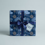 Square gift box in navy celestial wrapping paper with gold sun and moon designs, tied with a navy satin ribbon.