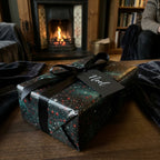 Elegant starry Christmas wrapping paper with black ribbon in front of a cozy fireplace by Wrapped Studios.