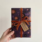 Celestial purple wrapping paper with gold suns and a velvet ribbon on a gift held by Wrapped Studios.