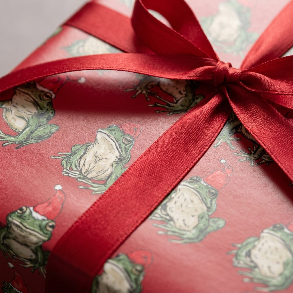 Cute frog Christmas wrapping paper by Wrapped Studios, featuring green frogs in Santa hats with a red ribbon.