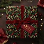 Square gift wrapped in festive Christmas paper with red berries, greenery pattern, and a velvet ribbon.