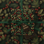 Elegant Christmas gift wrap with botanical leaf and red berry pattern, tied with a dark green ribbon and "For the Adventurer" tag.