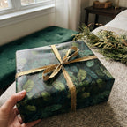 Hand holds a gift wrapped in festive evergreen Christmas gift wrap with gold ribbon.