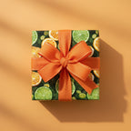 Square gift wrapped in citrus fruit & pine branch paper with a bright orange ribbon bow.
