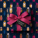 Navy gift box wrapped in colorful beetle print paper with bright pink ribbon and "Wrapped Studios" tag.