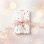 Playful polka dot birthday wrapping paper with a pink sheer ribbon and gift tag, set against a sparkly, light background.