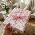 Pink cherry blossom floral wrapping paper with a ribbon bow by Wrapped Studios, held over a rustic wooden table.