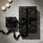 Elegant gifts wrapped in black floral wrapping paper with black ribbon, set on a marble surface with lit candles.
