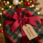 Plaid Christmas wrapping paper with a red bow and Wrapped Studios tag by a holiday tree.