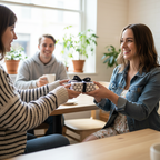 Two women exchanging a small, elegantly wrapped gift with a black bow at a cafe table, with a man smiling in the background.