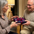 Senior couple exchanging a gift with floral wrapping paper and a purple ribbon in a cozy indoor setting.