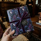 Hand holding a gift wrapped in gothic floral and raven paper with a luxury velvet purple ribbon by Wrapped Studios.