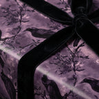 Elegant gift wrapped in purple wrapping paper with a crow and floral design, finished with a luxurious black velvet bow.