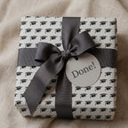 Graduation wrapping paper with black graduation caps, grey ribbon, and "Done!" tag by Wrapped Studios.