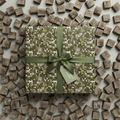 Modern gift wrapped in earthy green pixel pattern wrapping paper with ribbon and scattered wooden blocks.