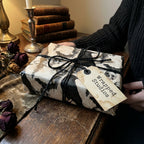 Goth wrapping paper with skulls and crows by Wrapped Studios, tied with black twine in a moody setting.