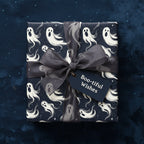 Square gift wrapped in ghost-patterned Halloween wrapping paper, with a dark grey ribbon and "Boo-tiful Wishes" tag.