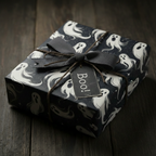Spooky Halloween gift wrapped in ghost-patterned wrapping paper, with a grey ribbon bow and "Boo!" tag, on dark wood.