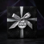 Black Halloween gift wrap with spiderwebs, silver ribbon, and "Creepy Delights" tag.