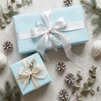 Light blue gift wrapping paper with white satin ribbon and winter greenery accents, ideal for Christmas presents.