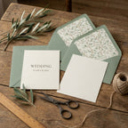 Sage green botanical wedding invitation suite on a rustic wood table with olive branches by Wrapped Studios.