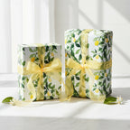 Two gifts wrapped in lemon floral wrapping paper with sheer yellow ribbon sit on a white table.