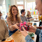 Friends exchanging a birthday gift wrapped in colorful candy print paper and a pink ribbon at a cafe.