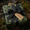 Green camouflage wrapping paper with a black bow and 'Mission Accomplished' tag by Wrapped Studios.