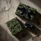 Green camouflage wrapping paper with black satin bows on rustic stone, by Wrapped Studios.