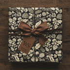 Square gift wrapped in elegant black and cream floral wrapping paper, tied with a brown ribbon bow.