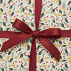 Holiday gift wrapped in botanical Christmas wrapping paper with red berry and leaf pattern, tied with a satin burgundy bow.