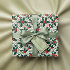Square gift wrapped in holly berry Christmas wrapping paper with sage green satin ribbon and "Happy Holidays" tag.