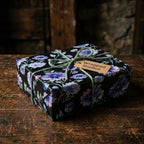 Luxury dark floral wrapping paper with purple flowers and green rope on a rustic table by Wrapped Studios.